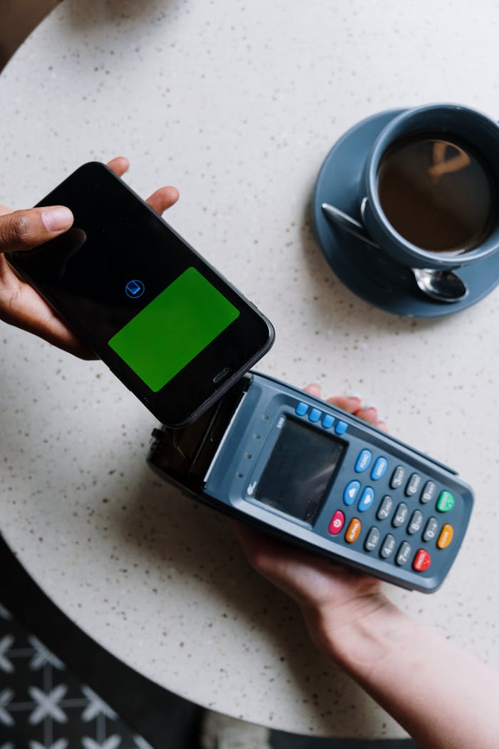 Smartphone making contactless payment on card reader next to coffee cup.