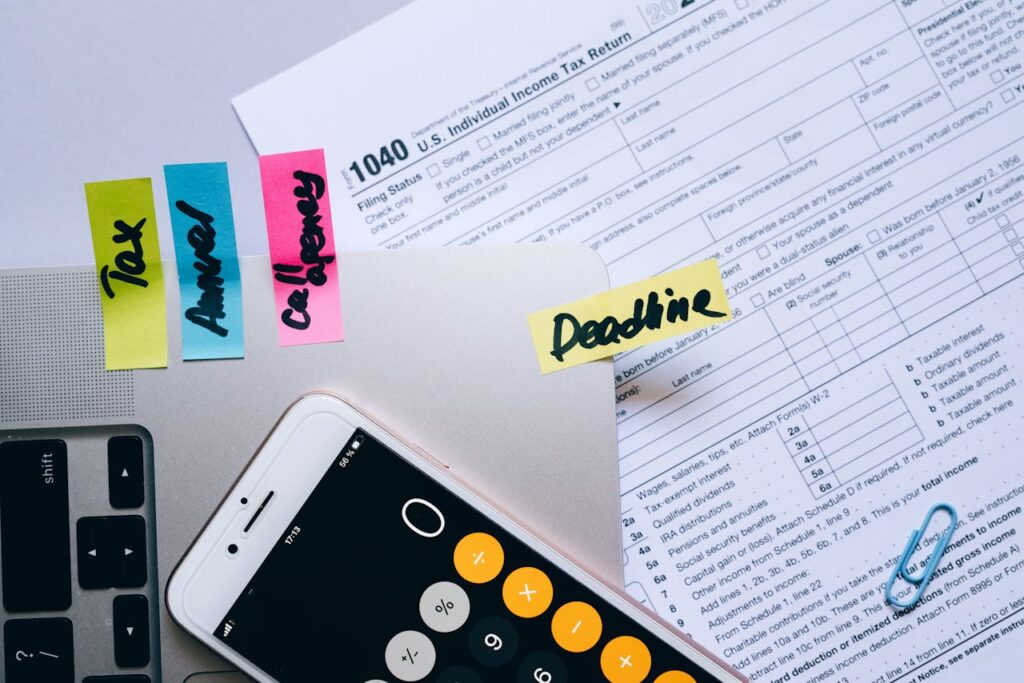 Office desk flat lay showing tax documents, calculator app on smartphone, sticky notes, and paperclips.